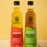 Amrutva Cold Pressed Groundnut and Sesame Oil Combo. This image shows two bottles of cold pressed oil; one groundnut and one sesame; alongside raw ingredients.